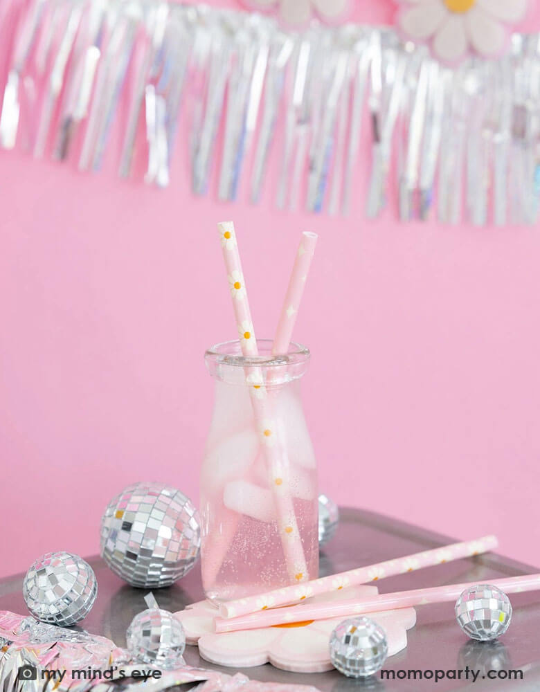 A milk bottle with pink soda and Momo Party's disco daisy reusable straws. Around the bottle there are Momo Party's disco daisy napkins and silver disco ball decorations. Above the the tableware is silver tinsel fringe garland, perfect for a groovy retro celebration!