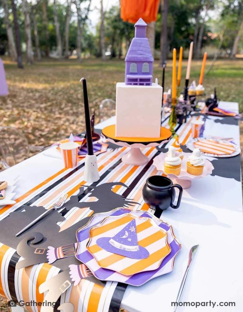 A Halloween witch themed party set up in a park featuring Momo Party's witch themed party supplies including purple and orange striped dinner plates, side plates and witch hat shaped napkins in the colors of purple, orange and peach. In the middle there's a haunted house shaped cake, some black and orange candles on ghost shaped candle stands on a black and orange striped table runner. Making this a great insp for kid's not-so-spooky Halloween party.