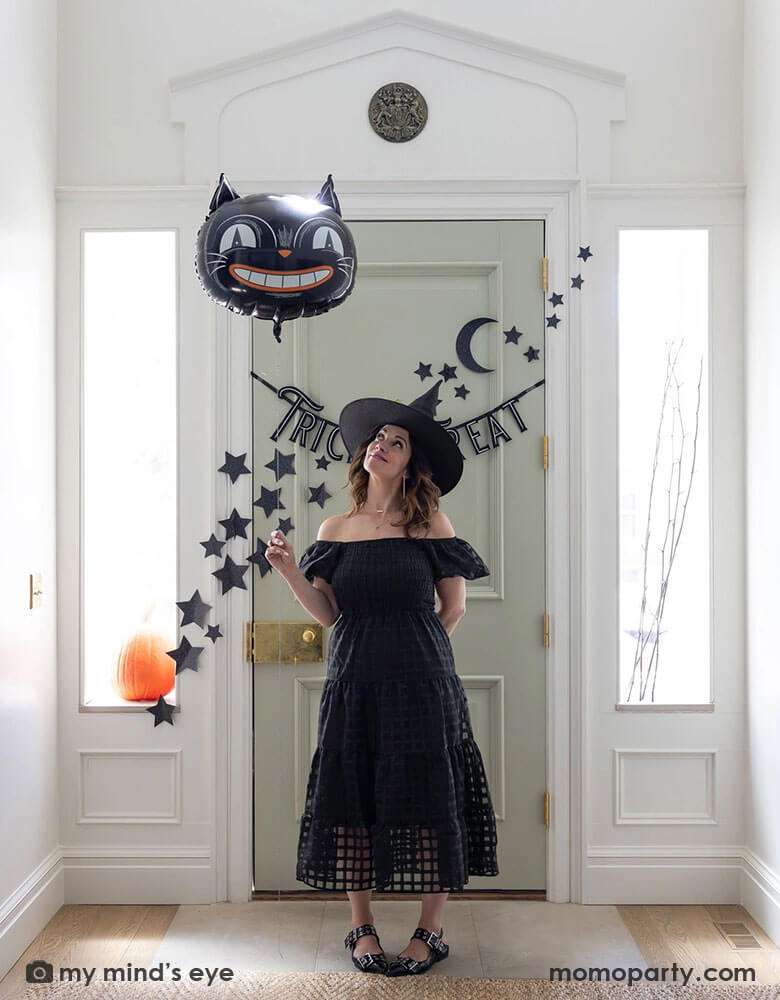 A lady dressed as a Halloween witch holding Momo Party's 24" black cat shaped foil balloon at the doorway decorated with Momo Party's 4ft Trick or Treat felt banner and glittered stars and moon wall paper decorations by My Mind's Eye. Behind the door through the side windows you can see a pumpkin and some spooky branches as the decorations, making this a perfect Halloween home decor idea for this spooky season!