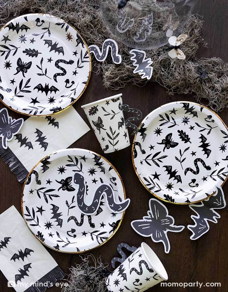 A table covered with Momo Party's spooky icon party supplies by My Mind's Eye including paper plates, cups, napkins and paper confetti. with some spooky dry grass around, this eerie line is perfect for a stylish Halloween party this spooky season.