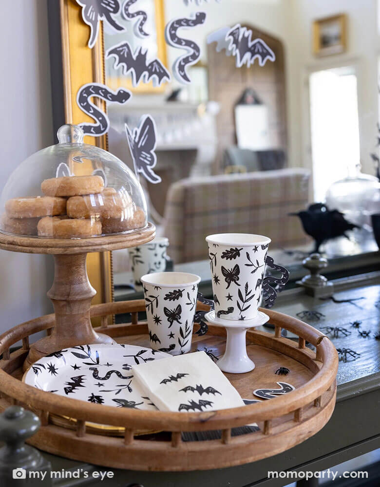 A spooky yet stylish Halloween table decoration featuring Momo Party's spooky icon party supplies collection including the snake handle party cups, the black and white spooky icon round plates and the bat guest napkins in a wooden tray which has some glazed donuts on a wooden cupcake stand. In the back there's a large mirror covered with Halloween paper decorations in black and white of Halloween elements including snakes, butterflies, bats, and butterflies. Making this a perfect stylish Halloween inspo.