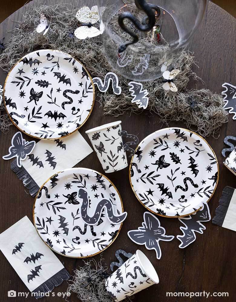 A table covered with Momo Party's spooky icon party supplies by My Mind's Eye including paper plates, cups, napkins and paper confetti. with some spooky dry grass around, this eerie line is perfect for a stylish Halloween party this spooky season.