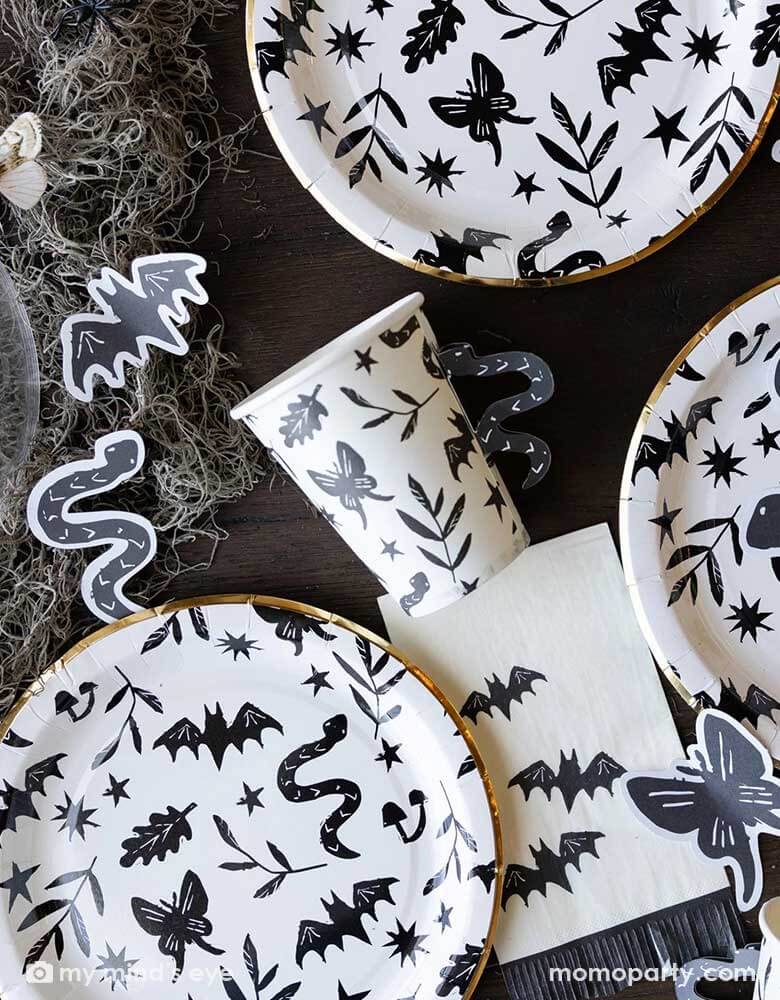 A table covered with Momo Party's spooky icon party supplies by My Mind's Eye including paper plates, cups, napkins and paper confetti. with some spooky dry grass around, this eerie line is perfect for a stylish Halloween party this spooky season.