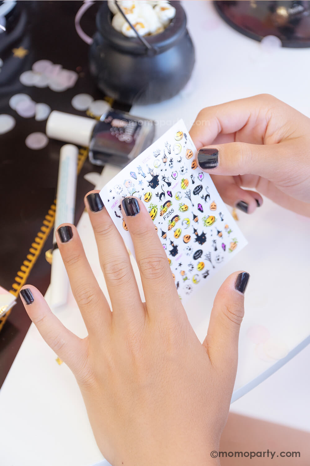 A girl wears the black nail polish in a witch themed party table looking at the nail sticker sheet with Halloween themed nail stickers including black cats, haunted mansions, Jack-o-lantern, crescent moon, and skulls from the Snails Nail Polish Halloween Spooky Pack - which makes a perfect party favor or activity for kid's Halloween party.