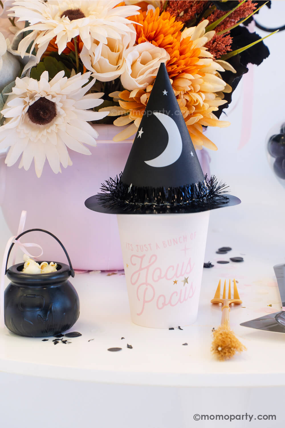 A witch themed Halloween party table styled by Momo Party featuring pink Halloween party supplies including the pink Hocus Pocus party cup which is topped with the witch party hat with a crescent moon on it. On the table there's a broom shaped fork next to a witch hat shaped plate and a mini cauldron filled with popcorn. With the fall flower arrangement in the pink cauldron in the back and black confetti around the table, it makes a spooky cute tablescape inspo for a girly Halloween bash this spooky season