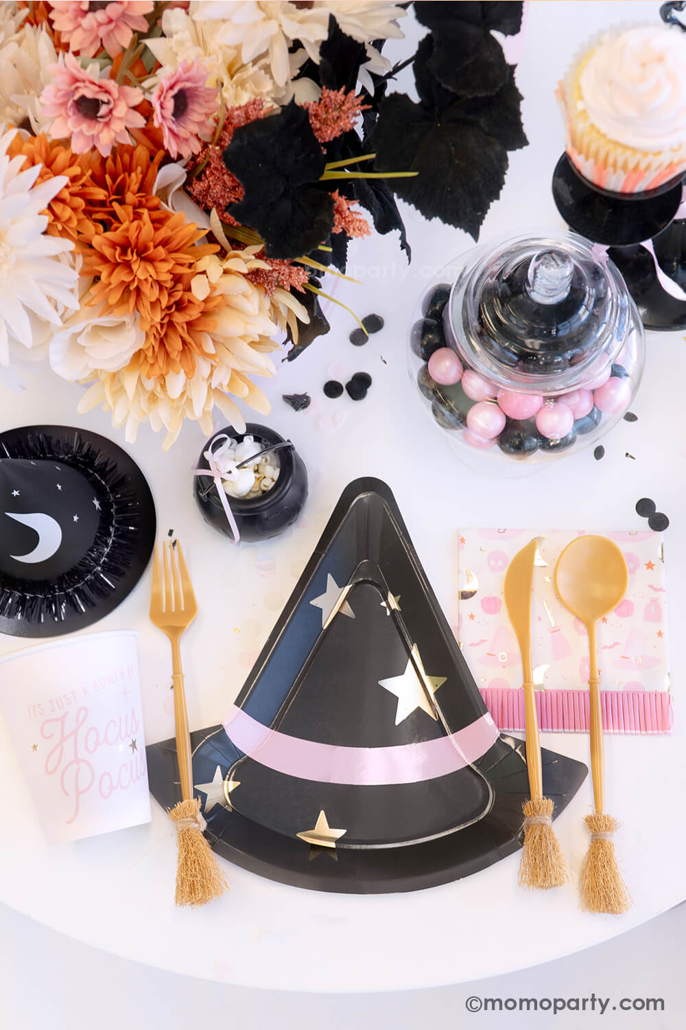 A witch themed Halloween party table styled by Momo Party featuring pink Halloween party supplies including the  pink witch hat shaped plate, witch's broom cutlery set, Hocus Pocus party cups and napkins, and the witch party hat. On the table, there's mini cauldron with popcorn, some pink and black gum balls and a fall flower arrangement in the pink cauldron - making it a spooky cute tablescape inspo for a girly Halloween bash this spooky season.