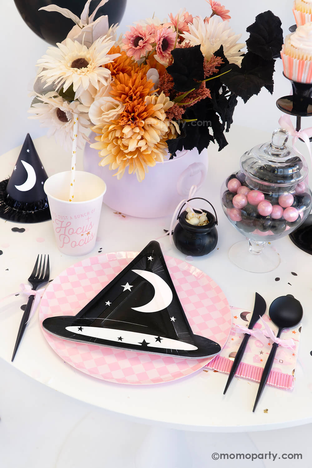 A witch themed Halloween party table styled by Momo Party featuring pink Halloween party supplies including the  pink Hocus Pocus party cup, napkin, witch hat shaped plate on a pink checkered plate, and a witch party hat. On the table, there's mini cauldron with popcorn, some pink and black gum balls and a fall flower arrangement in the pink cauldron - making it a spooky cute tablescape inspo for a girly Halloween bash this spooky season.