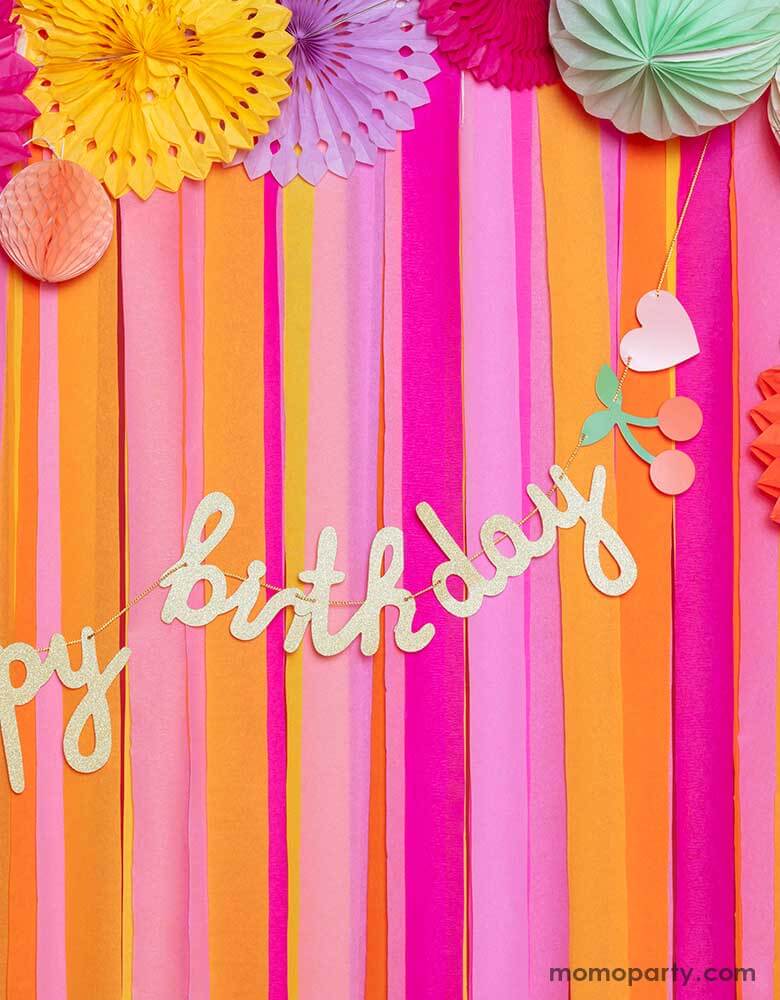 A closeup of a pink room decorated with colorful paper fans and streamers adorn with happy birthday banner with rainbows and cherries. A cheerful and festive decorations for birthdays of any age.