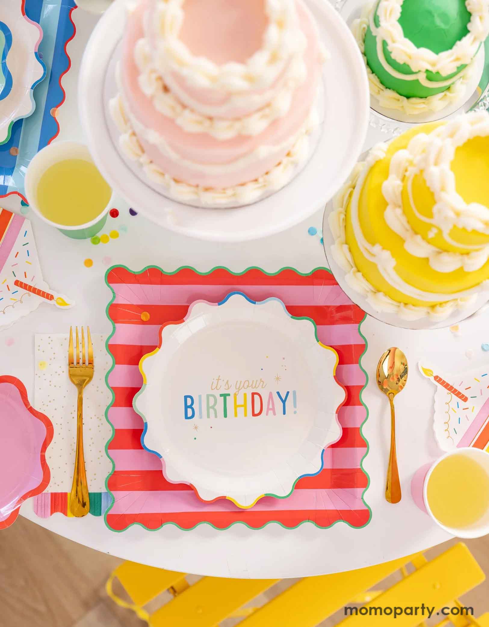 A festive and cheerful birthday party table featuring Momo Party's Happy Birthday party tablewares including the 9" It's Your Birthday Plates, the red striped dinner plates, the slice of cake shaped napkins, and the rainbow fringe guest towels by My Mind's Eye. On the table there are multiple birthday cakes in pastel rainbow colors, making this a modern and adorable table setting for a happy birthday celebration for kids of any age!