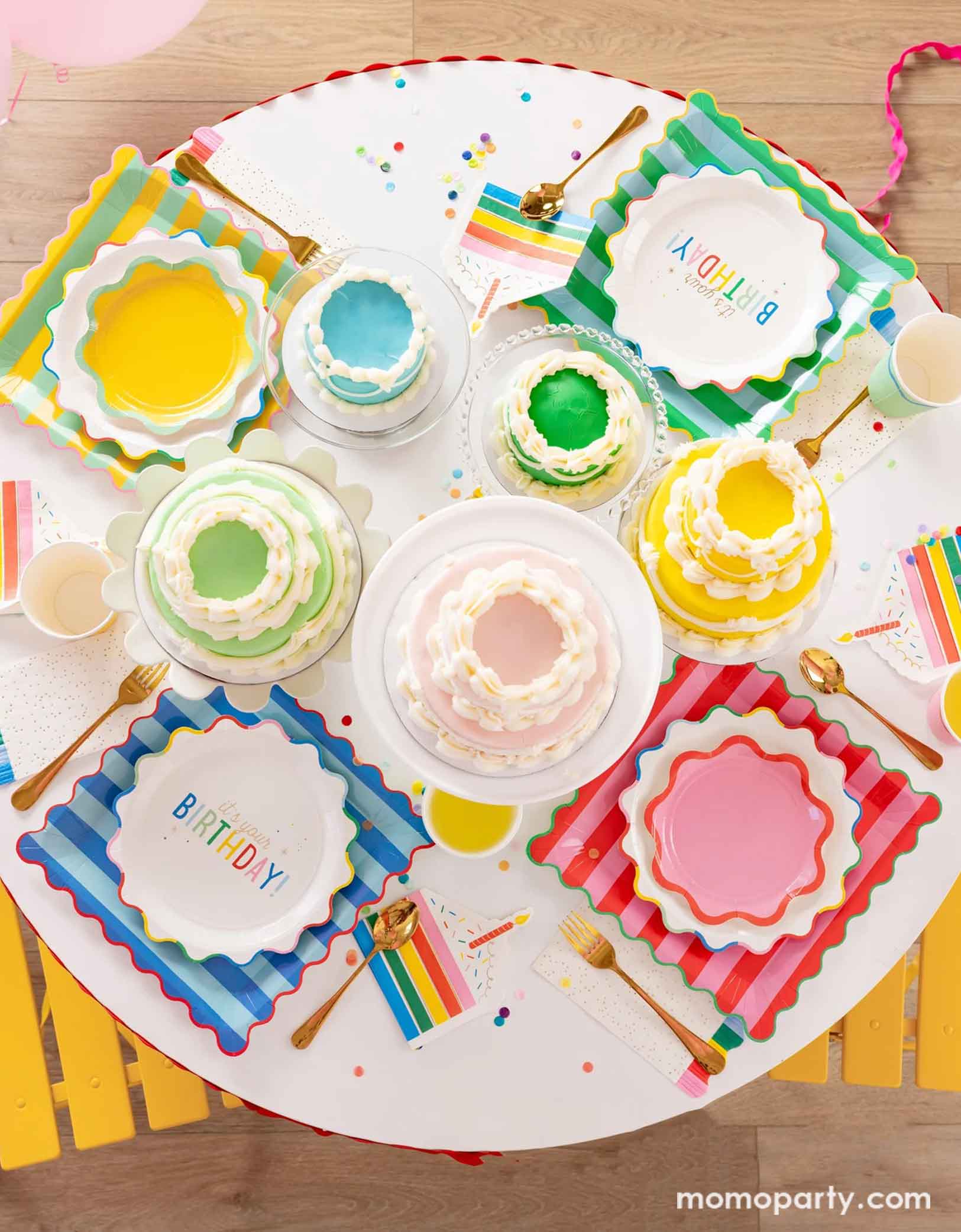 A overhead shot of a festive and cheerful round birthday party table featuring Momo Party's Happy Birthday party tablewares including the 9" It's Your Birthday Plates, the striped dinner plates in four different colors the ric rac edge dessert plates, the slice of cake shaped napkins, and the rainbow fringe guest towels. On the table there are multiple birthday cakes in pastel rainbow colors in different sizes, making this a modern table setting for a happy birthday celebration for kids of any age!