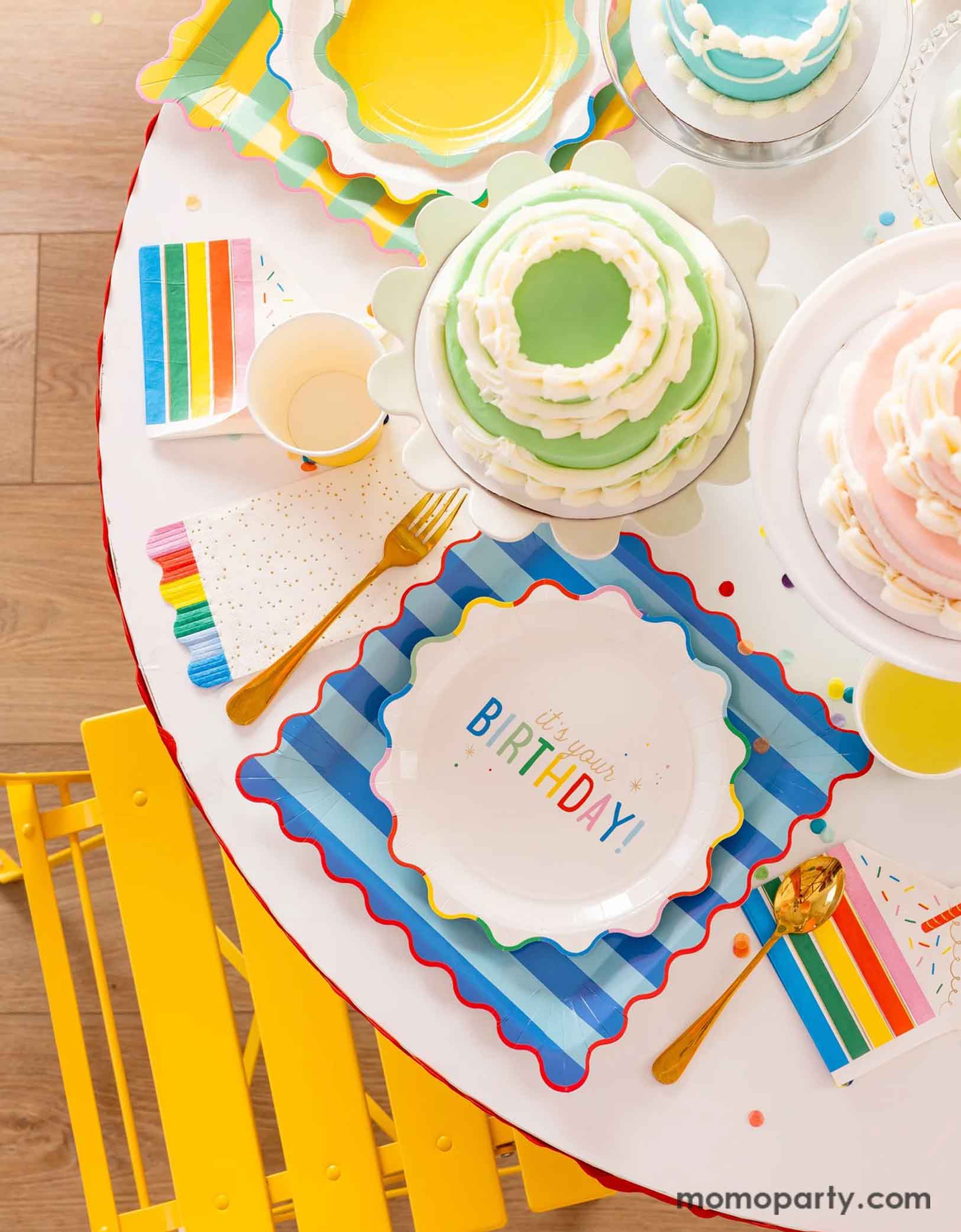 A festive and cheerful birthday party table featuring Momo Party's Happy Birthday party tablewares including the 9" It's Your Birthday Plates, the striped dinner plates, the slice of cake shaped napkins, the rainbow fringe guest towels by My Mind's Eye. On the table there are multiple birthday cakes in pastel rainbow colors, making this a modern and adorable table setting for a happy birthday celebration for kids of any age!