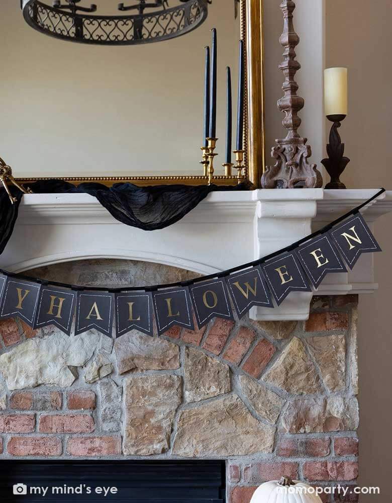 A close up shot of Momo Party's Happy Halloween banner hung over a mantel that's decorated with elegant Halloween decorations including a black and gold chandelier, black and gold candle holders, a beige colored pumpkin and a gold framed vintage mirror.