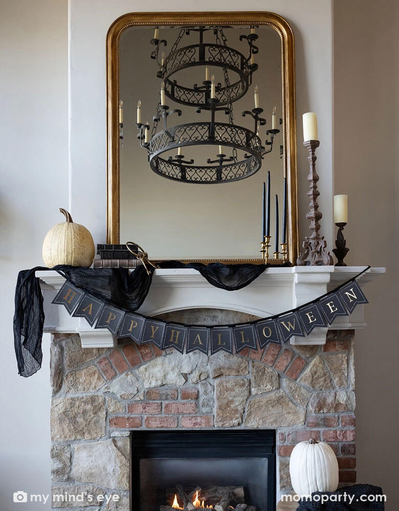 A Halloween fireplace mantel decorated with some elegant Halloween decorations including a black and gold chandelier, black and gold candle holders, some white and beige colored pumpkins and a gold framed vintage mirror. In the middle hung Momo Party's black and gold Happy Halloween banner by My Mind's Eye, making this a perfect inspo for a chic and elegant Halloween home decor.