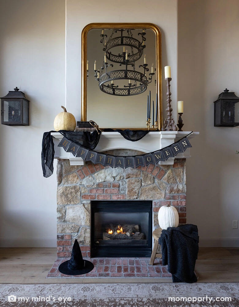 A Halloween fireplace mantel decorated with some elegant Halloween decorations including a black and gold chandelier, black and gold candle holders, some white and beige colored pumpkins, a witch hat and a gold framed vintage mirror. In the middle hung Momo Party's black and gold Happy Halloween banner by My Mind's Eye, making this a perfect inspo for a chic and elegant Halloween home decor.