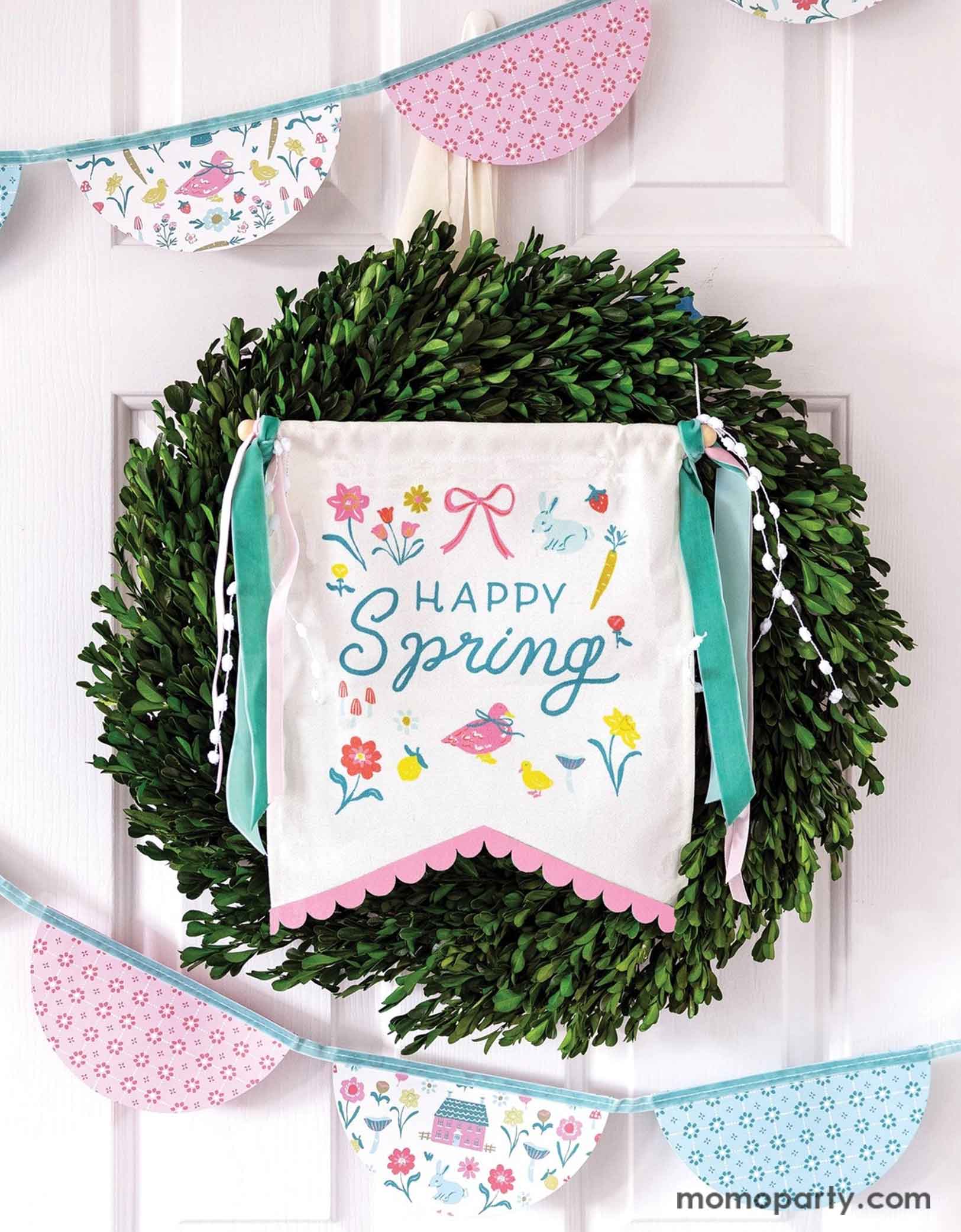 A white house door decorated with a wreath and Momo Party's 10.5" x 15" Happy Spring Canvas Banner by My Mind's Eye, featuring a playful "happy" message. With spring inspired illustrations like birds, flowers, bunnies, carrots and bows in pastel colors. Around the wreath there's a spring inspired bunting garland banner in pastel color and spring illustration motif, a prefect way to welcome a fresh and happy season of spring.