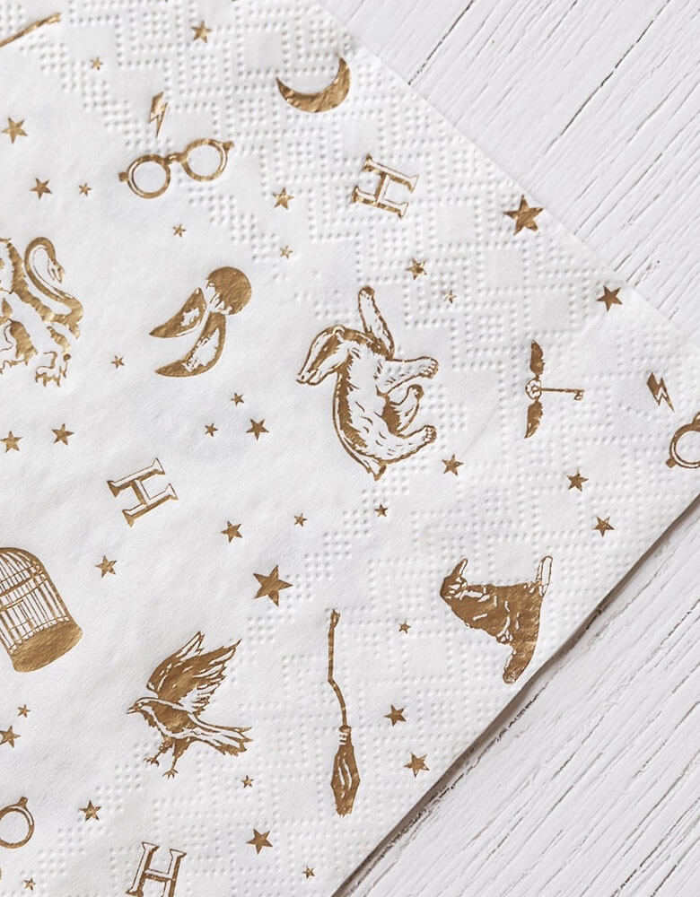 A close up shot of Momo Party's 6.5" x 6.5" Harry Potter Large Napkins by Bonjour Fete. Comes in a set of 24 napkins with elements including glasses, wizard hats, moons, stars and owls from Harry Potter in gold accent. Throw a magical bash with these Harry Potter party napkins. With whimsical gold accents, this modern design is perfect for any wizarding event.