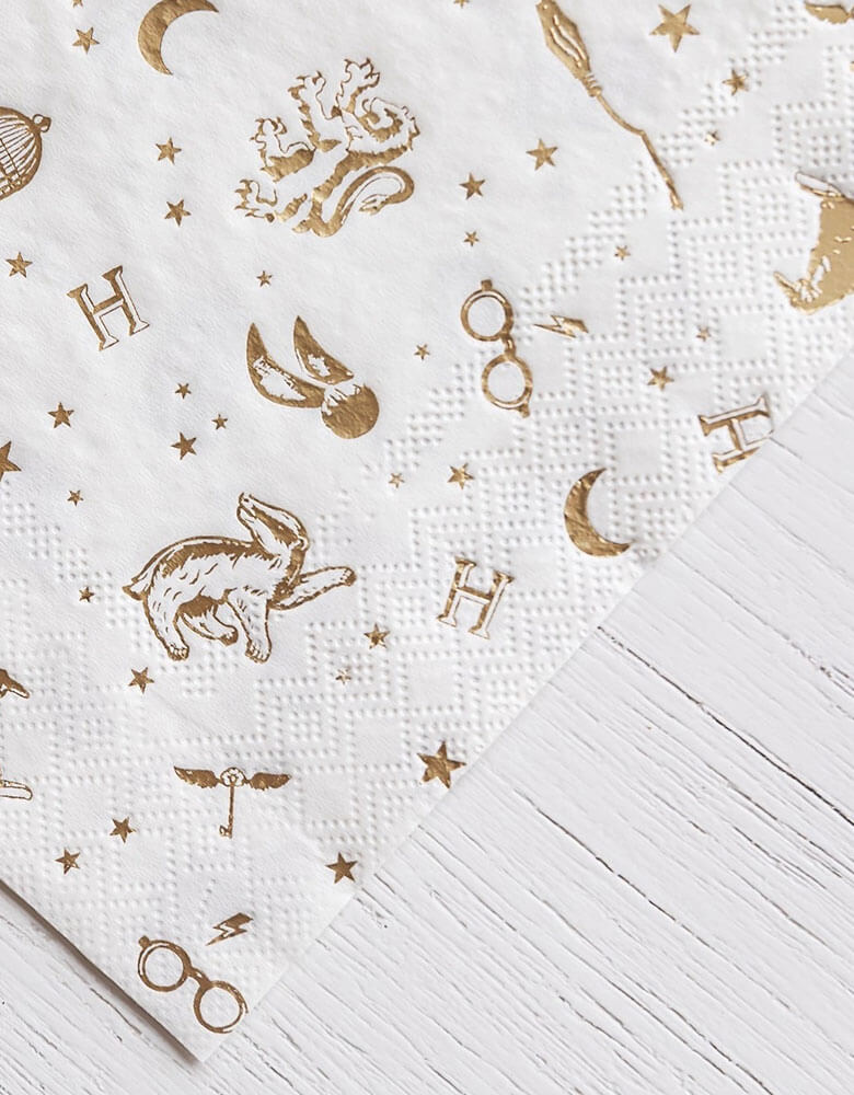 A close up shot of Momo Party's 6.5" x 6.5" Harry Potter Large Napkins by Bonjour Fete. Comes in a set of 24 napkins with elements including glasses, wizard hats, moons, stars and owls from Harry Potter in gold accent. Throw a magical bash with these Harry Potter party napkins. With whimsical gold accents, this modern design is perfect for any wizarding event.