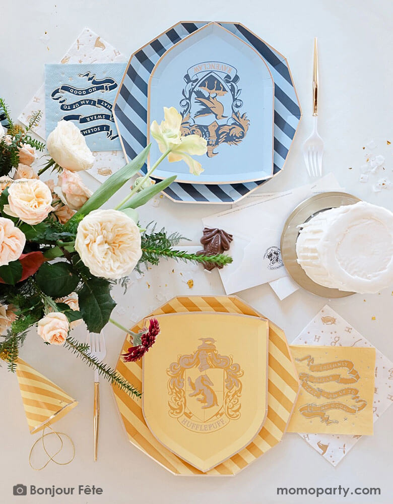 A beautiful and elegant Harry Potter themed party tablescape features Momo Party's Harry Potter house pride shield-shaped 7" side plates with Hufflepuff and Ravenclaw logos and colors paired with 10.25 striped dinner plates. Next to the plates are Harry Potter motif large napkins in gold foil and matching small napkins. On the table there's a candle stick shaped buttercream cake, a letter from Hogwarts, some gold confetti and party hats, along with a beautiful flower bouquet.