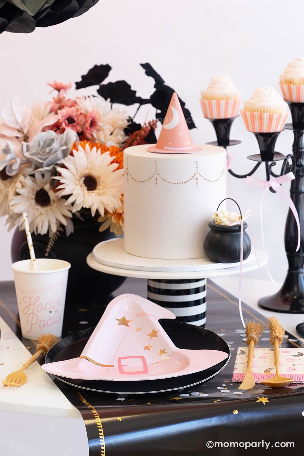 An adorable witch-themed pink Halloween party table setting featuring various pink Halloween party tableware from Momo Party including the pink witch hat shaped plate, Hocus Pocus small napkins, party cups and witch broom cutlery set on the mystic table runner. On the table there's a pink fondant cake adorned with a pink witch hat as the topper and a mini cauldron tied with a pink ribbon. With the pink boho floral arrangement and pink cupcakes on the vintage black candle holder a spooky cute table is ready!