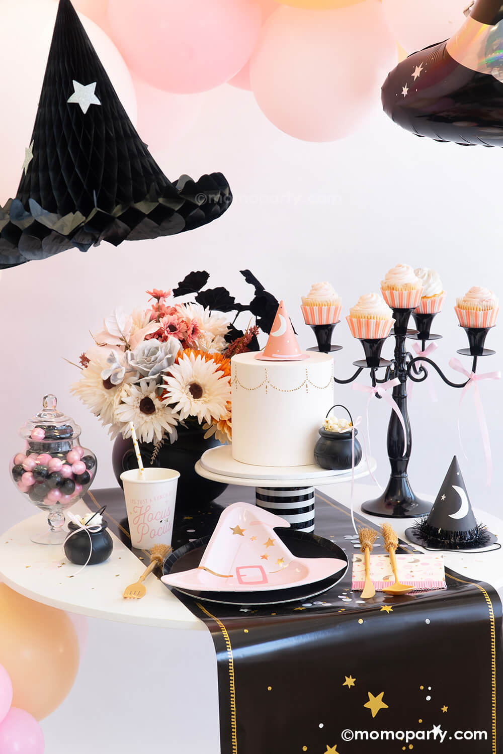 An adorable witch-themed pink Halloween party table setting featuring various pink Halloween party tableware from Momo Party including the pink witch hat shaped plate, Hocus Pocus small napkins, party cups and witch broom cutlery set on the mystic table runner. On the table there's a pink fondant cake adorned with a pink witch hat as the topper and a mini cauldron tied with a pink ribbon. With the pink boho floral arrangement and pink cupcakes on the vintage black candle holder a spooky cute table is ready!