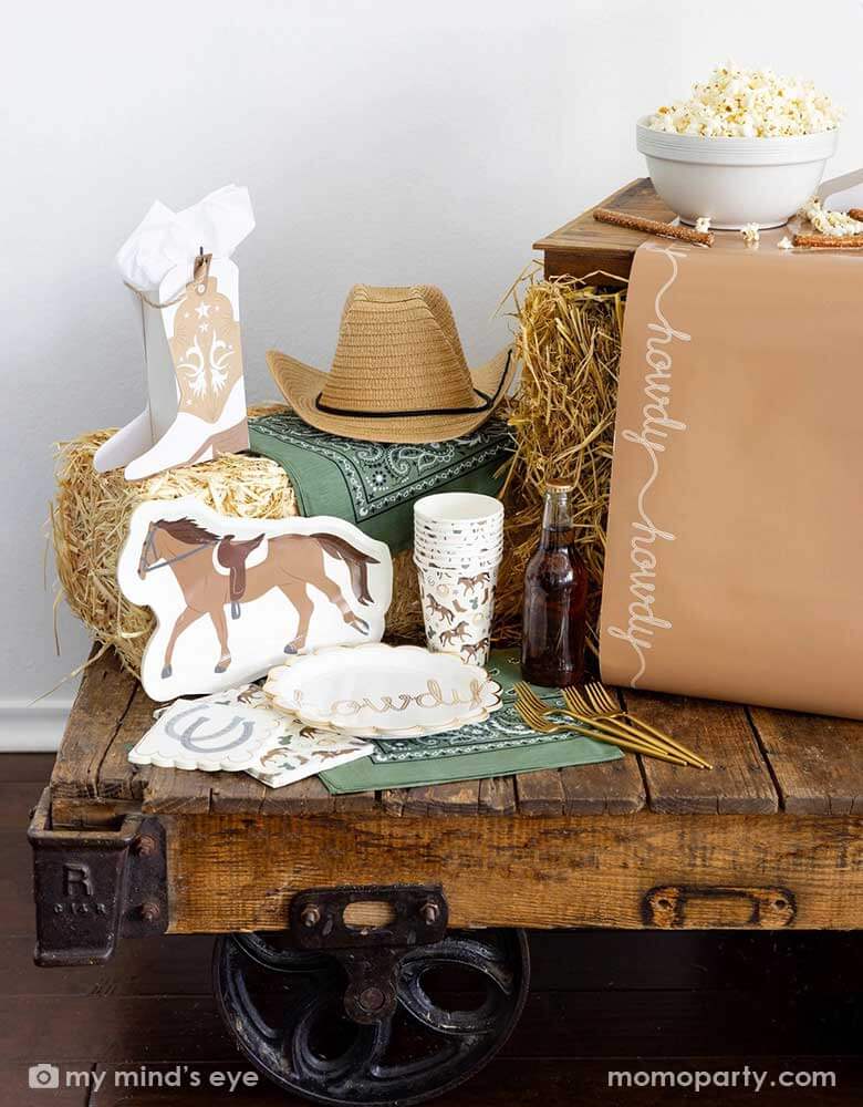 A cowboy themed party table featuring Momo Party's cowboy themed party supplies including horse shaped plates, cowboy patterned cups, napkins, table runner and cowboy boot shaped treat boxes. On the table there's a cowboy hat, straw bale, and a green bandana as decoration, perfect for a Western rodeo themed celebration, be a First Rodeo boy's first birthday party or a fun cowboy bash!