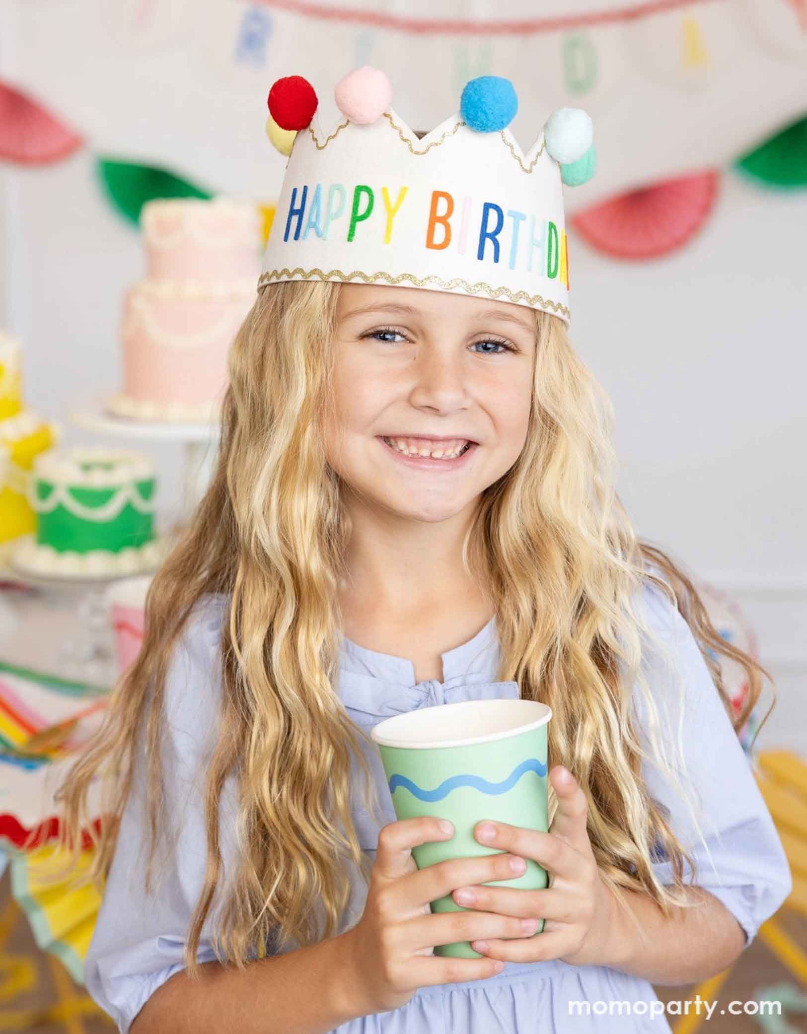 A school-aged girl wearing a colorful felt happy birthday crown with pom poms holding Momo Party's 12 oz green Ric Rac party cup in her hands. In the back there's a table full up birthday cakes in different pastel colors and some festive happy birthday banners on the wall, making this a charming scene for kid's birthday party celebration.