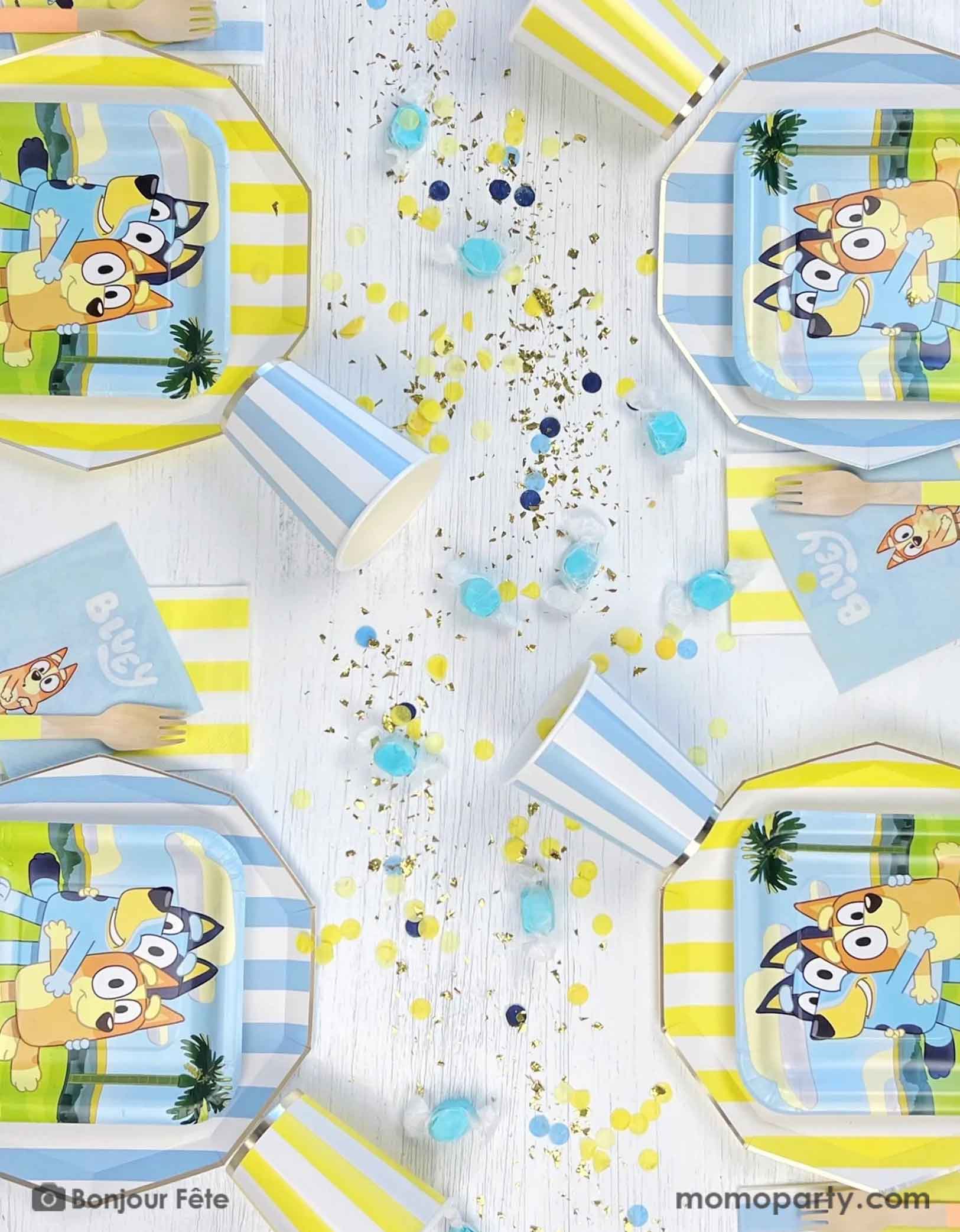 An overhead shot of a Bluey themed kid's birthday party table featuring Momo Party's modern blue and yellow cabana striped tablewares including paper plates, paper cups and napkins. On the dinner plates there are square side plates and party napkins featuring Bluey charcters including Bluey and Bingo, with matching colored confetti and blue colored candies on the table. 