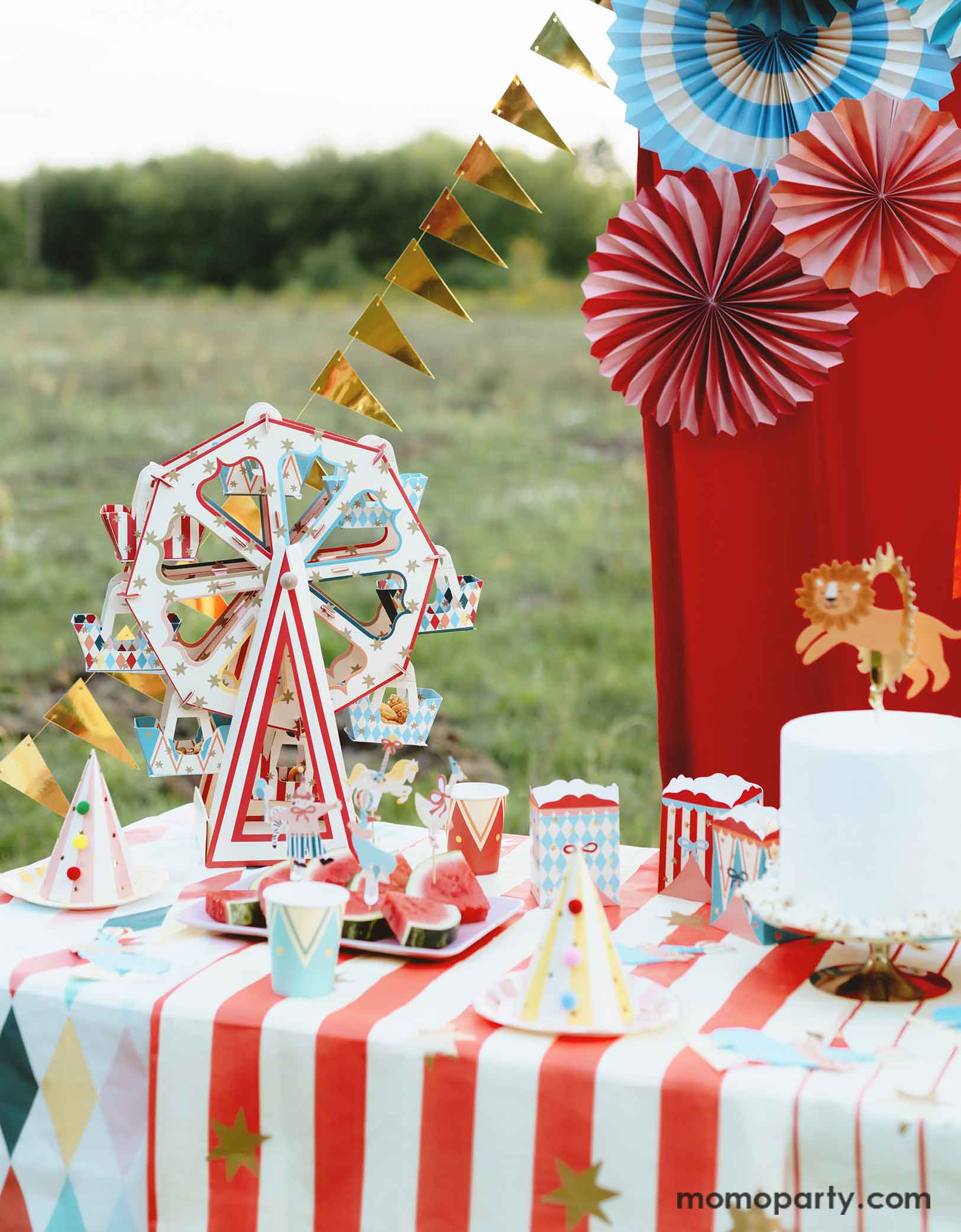 An adorable circus & carnival themed birthday party table set up and decorations feature various circus themed party supplies from Momo Party, including a stunning ferris wheel treat stand centerpiece with its circus-inspired design is loaded with cupcakes, party plates, hats, cups, treat boxes and a birthday cake on a carnival themed tablecloth. In the back there's a red stage curtain as a party backdrop decorated with circus themed birthday banner with circus animal party pennants and colorful party fans.