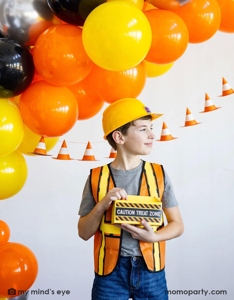 A school aged boy in his construction costume and hard hat holding a toolbox shaped treat box standing in front of a festive construction themed party decoration which features a balloon garland in black, orange and yellow and a construction cone banner hung over on the wall - a perfect decoration idea for kid's construction builder themed birthday celebration party.