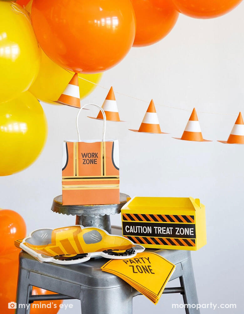 A festive kid's construction themed birthday party decoration set up featuring various construction party supplies and decorations from Momo Party including dumper truck shaped plates, party zone napkins, work zone vest shaped party bags, construction toolbox shaped treat boxes by My Mind's Eye. In the back there's a balloon garland in classic construction colors of orange and yellow, adorned with a construction cone paper banner, making this a perfect inspo for boy's construction birthday party!