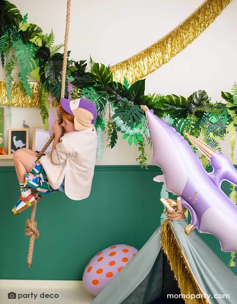 A boy hanging on a rope in a room decorated with dinosaur party decorations including a tropical leaves garland, a gold tinsel fringe garland, a lilac PTERODACTYL shaped foil balloon - making this a fun and modern set up for kid's dinosaur themed birthday parties.