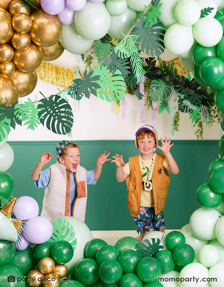 Two school aged boys in a tropical dinosaur themed balloon decoration set up featuring balloon garland in different shades of green, lilac and chrome gold. The organic balloon garland is adorned with Momo Party's tropical leaves paper decorations by Party Deco. Along with the gold tinsel fringe garland, it creates an amazing prehistorical vibe, a perfect inspo for kid's dinosaur themed birthday party or a tropical themed bash in summer!