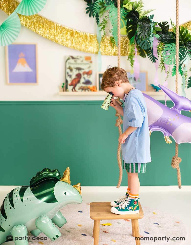 A boy is watching Momo Party's Triceratops shaped foil balloon through his binoculars. He's in a room decorated with prehistorical  themed decoration including a lilac PTERODACTYL shaped balloon, a gold tinsel fringe garland, a tropical leaves garland from Momo Party.