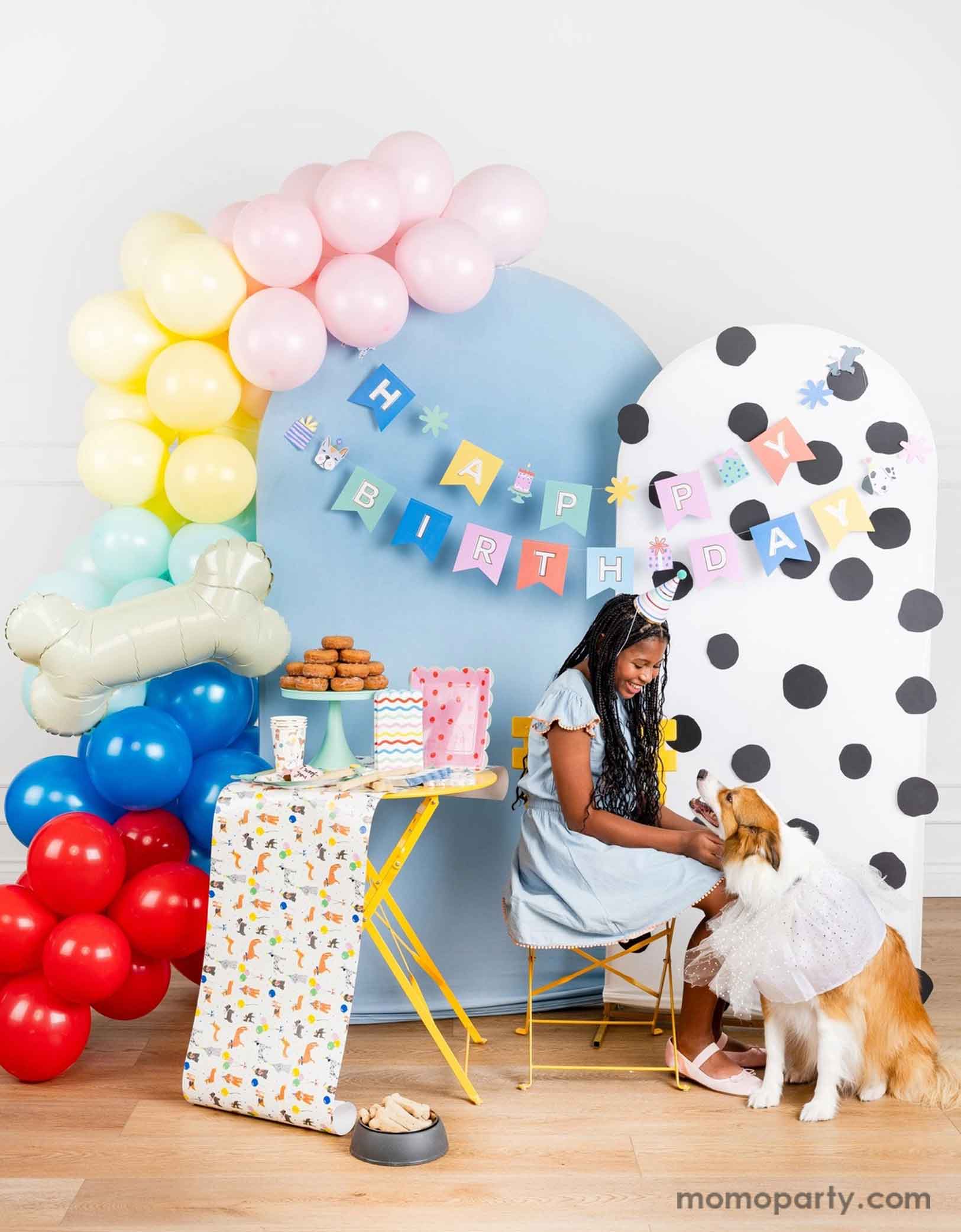 A dog themed party decoration set up featuring Momo Party's dog themed birthday supplies by My Mind's Eye. Including dog Happy Birthday party banner hung over a polka dotted and a pastel blue party backdrop with a pastel colored party balloon garland and a bone shaped foil balloon. On the table there are some dog themed party tablewares sugar cookies, treats and a donut tower. A girl in a birthday hat is sitting on the side and petting her dog happily celebrating her birthday.