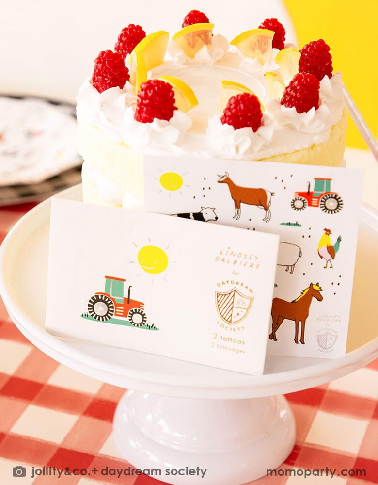 Momo Party's on the farm temporary tattoos and stickers sheets standing next to a buttercream birthday cake adorned with lemon slices and raspberries on a cake stand on a red gingham tablecloth in a kid's farm themed birthday party.