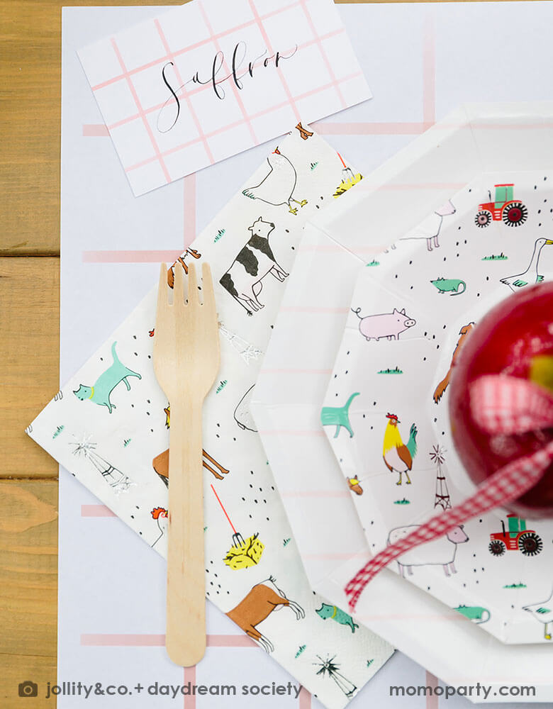 A farm themed kid's birthday party placeset featuring Momo Party's On the farm party plate & napkin paried with pink striped plates and placemat. On the plates there is a candy dipped apple adorned with red gingham ribbons. 