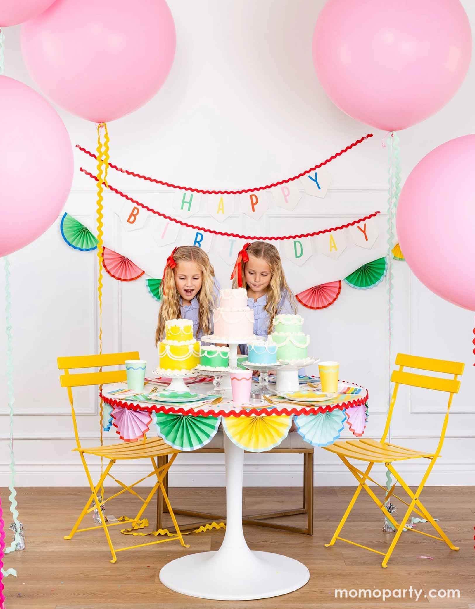 A cheerful and colorful birthday party set up featuring various festive birthday party supplies from Momo Party including rainbow colored tablewares of plates, cups, napkins on the table along with multiple pastel colored birthday cakes. The table is decorated with colorful bunting garland around and in the back of the wall hung a Happy Birthday felt garland and the colorful bunting banner. On the sides are some jumbo pink balloons floating above and two girls standing excitedly next to the cakes.