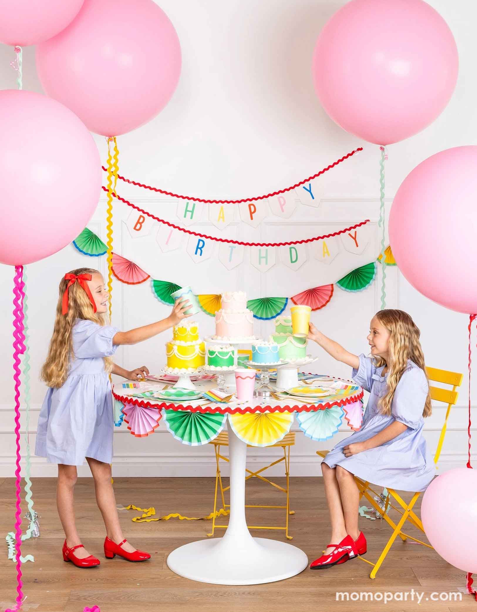 A cheerful and colorful birthday party set up featuring various festive birthday party supplies from Momo Party including rainbow colored tablewares of plates, cups, napkins on the table along with multiple pastel colored birthday cakes. The table is decorated with colorful bunting garland around and in the back of the wall hung a Happy Birthday felt garland and the colorful bunting banner. On the sides are some jumbo pink balloons floating above and two girls  sitting at the table and holding cups.