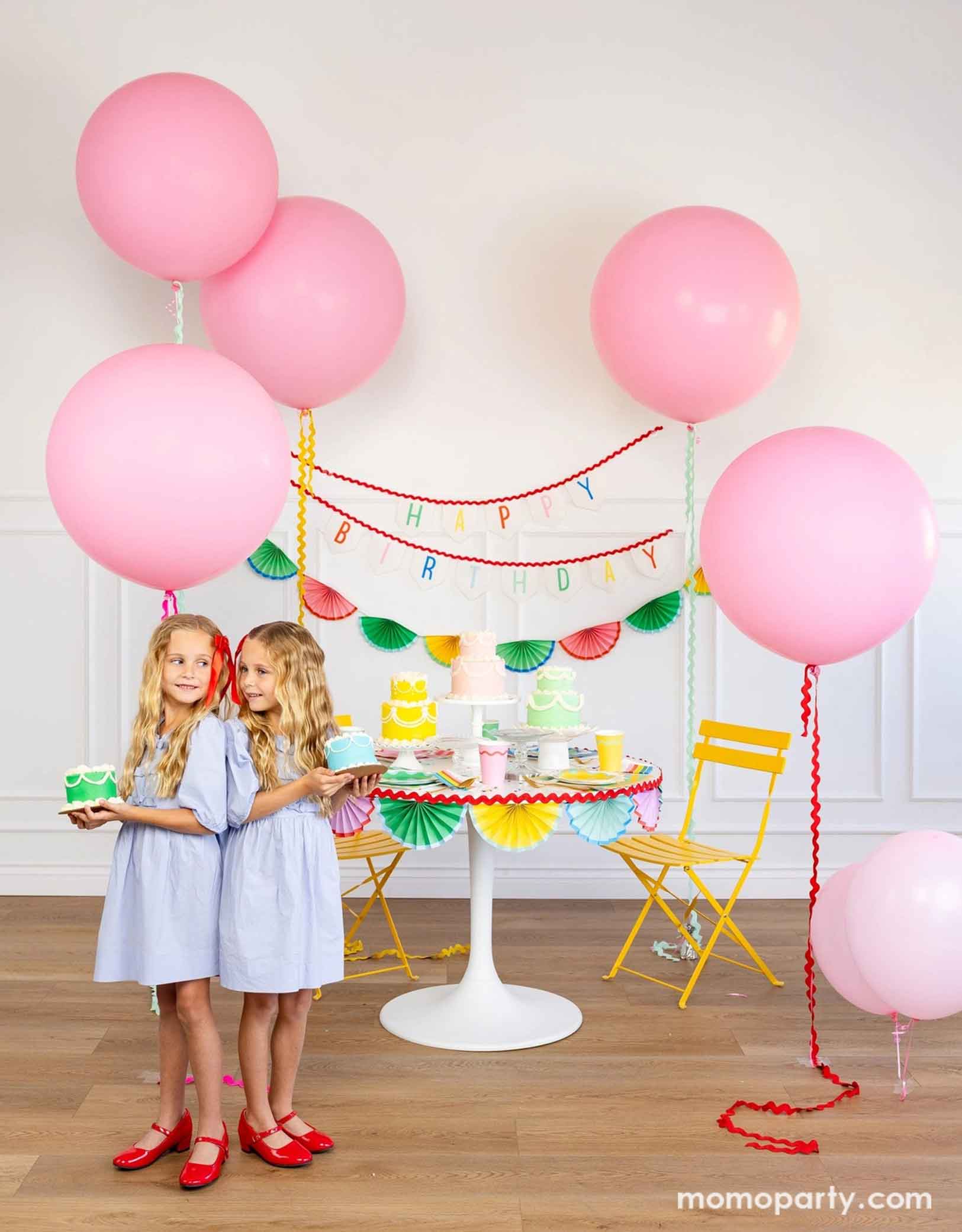 A cheerful and colorful birthday party set up featuring various festive birthday party supplies from Momo Party including rainbow colored tablewares of plates, cups, napkins on the table along with multiple pastel colored birthday cakes. The table is decorated with colorful bunting garland around and in the back of the wall hung a Happy Birthday felt garland and the colorful bunting banner. On the sides are some jumbo pink balloons floating above and two girls standing in the front holding birthday cakes.