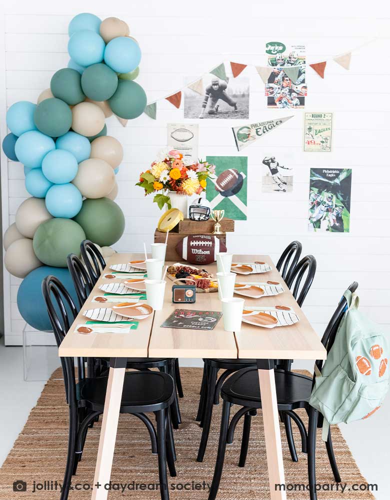 A kid's football birthday party set up with a modern party table filled with football themed tableware from Momo Party including the die-cut football shaped plates, black striped plates, green and mint napkins and party cups. In the center of the table there's a cheese charcuterie board and some decorations like a football, a trophy and a football helmet as the centerpiece. On the wall there are some old school football party banners, posters and party balloons to add some festivities to the celebration!