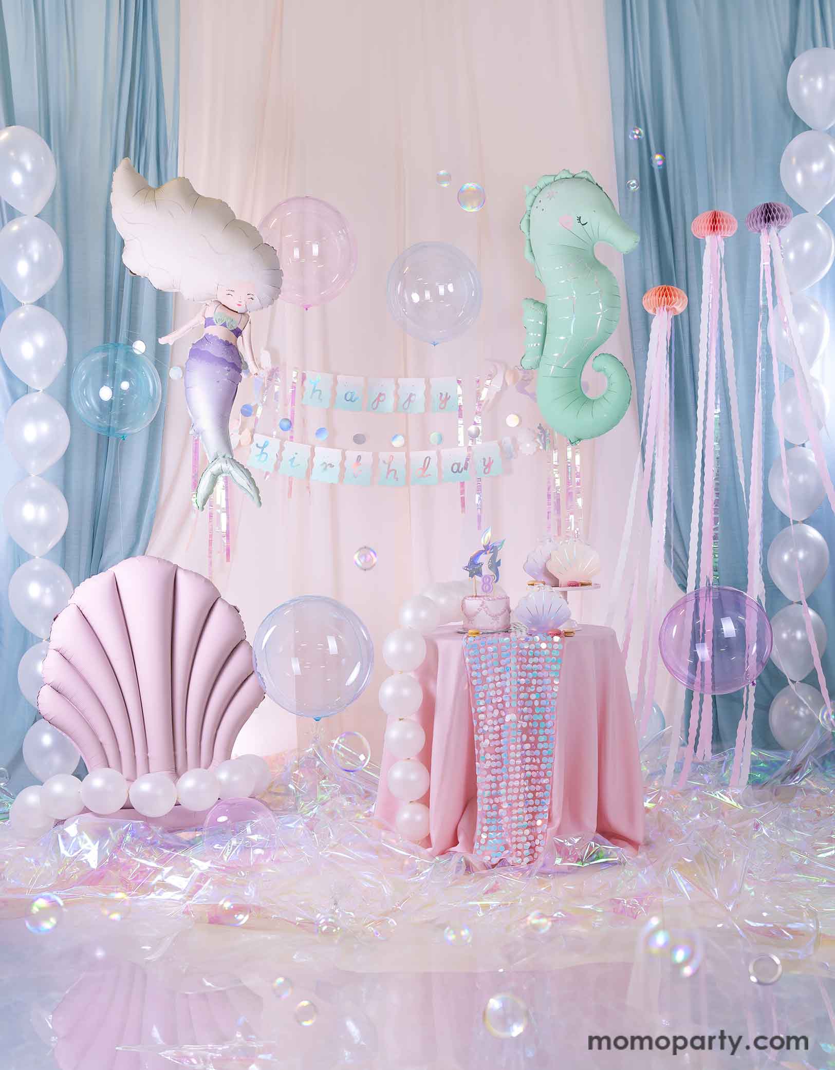 An enchanting and dreamy kid's mermaid party set up featuring Momo Party's under the sea mermaid themed party supplies including a floating mint seahorse balloon, a mermaid shaped balloon, a lilac seashell standing foil balloon adorn with pearl white balloons, the mermaid happy birthday part garland, and a table with mermaid themed tableware. With a white and light blue curtain in the back as the backdrop, lots of see-through bubble balloons and an iridescent cellophane on the floor.