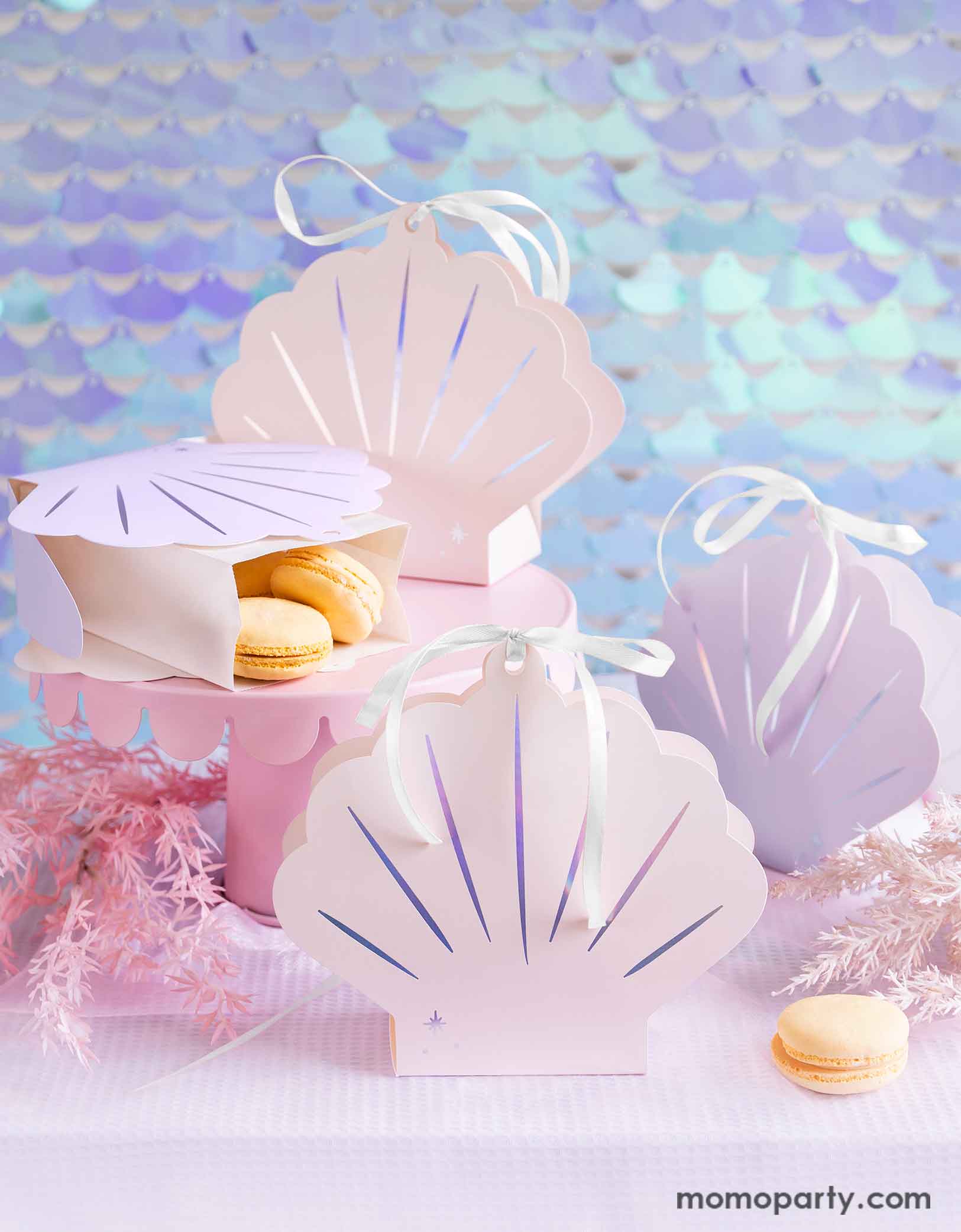 A mermaid birthday party table featuring Momo Party's 6.3" x 6.9" x 2.2" Seashell Treat Boxes by Party Deco. These boxes come in a set of 4 boxes in 2 colors of ombre pink and ombre lilac with a bow tied at the top. In the boxes there are some vanilla macarons on a pink cake stand surrounded by pink seagrass decorations. Behind the table there's a seashell-shaped iridescent sequin curtain as the backdrop, creating a dreamy and whimsical scene for a magical under the sea bash. 