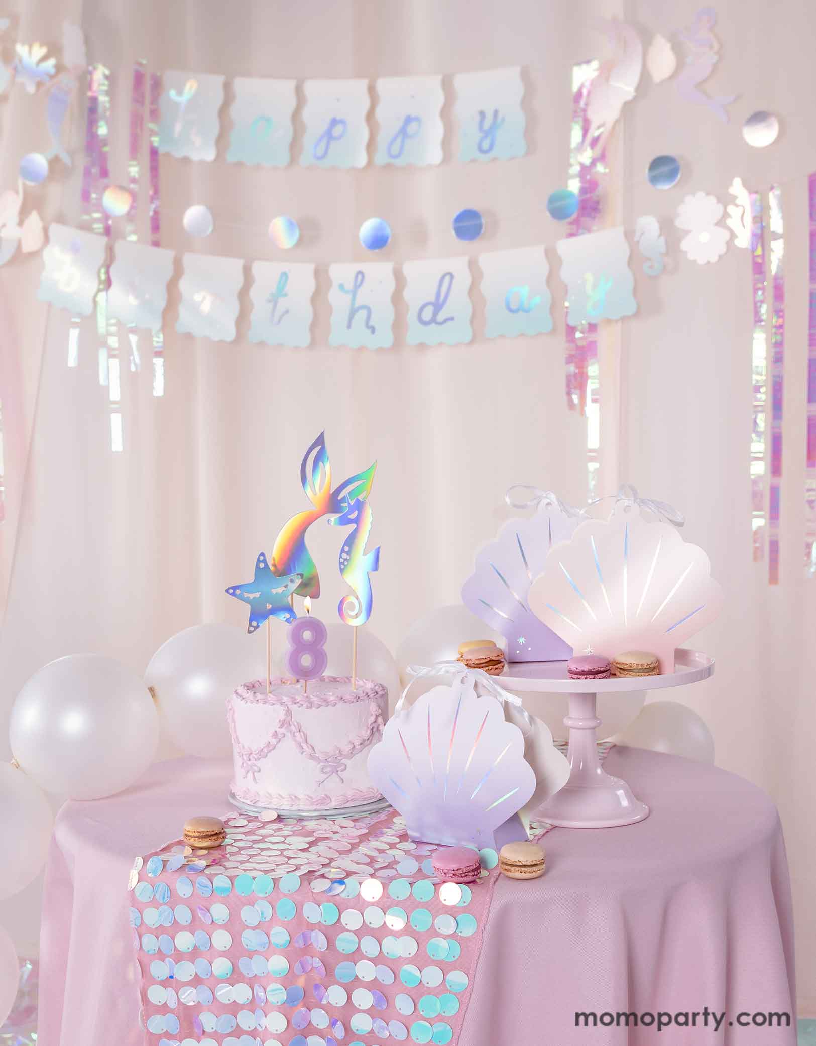 A mermaid birthday party table set uo featuring various mermaid themed party supplies from Momo Party including the  ombre pink and lilac shell shaped treat boxes, a pink butter cream cake topped with mermaid themed toppers. Behind the table there's a pink curtain adorned with the mermaid happy birthday party banner as the backdrop. Along with pearl white balloons around, creating a dreamy and whimsical scene for a magical under the sea bash. 