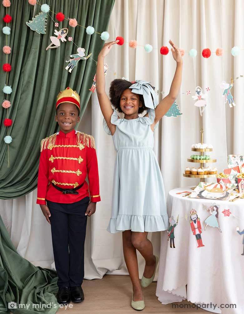 A girl and a boy dressed as the Nutcracker characters in a Nutcracker themed holiday party. The girl is doing a ballet dance pose and they're standing next to a party table filled with Holiday treats and Nutcracker themed party supplies. In their back there's a stage curtain in satin green and beige which is decorated with Momo Party's Nutcracker 6' garland with pom-poms which adds a festive touch to the Holiday decoration.