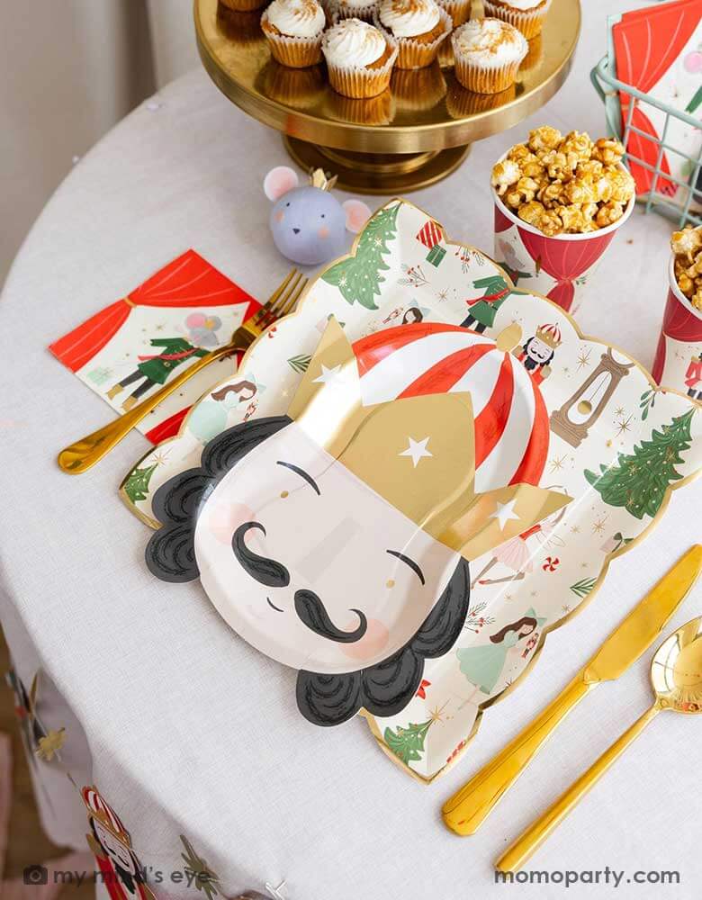 A festive Holiday party table featuring Momo Party's Nutcracker themed tableware including the Nutcracker pattern plate, king head shaped plate, Nutcracker small napkins, Nutcracker party cups and the Nutcracker mouse surprise ball by My Mind's Eye. On the party table there are also some kid's friendly treats including cupcakes and popcorns - making this a perfect inspo for kid's Nutcracker themed Holiday celebration this Christmas.