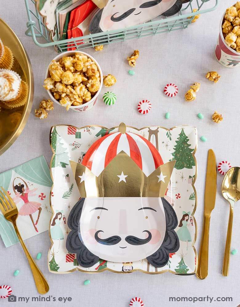 A festive Nutcracker themed Holiday party tablescape featuring Momo Party's Nutcracker party supplies including the Nutcracker pattern plate, Nutcracker head shaped plate, Nutcracker napkins, party cups which are filled with popcorn. On the table there are some Holiday treats including  green and red candies, peppermint and cupcakes making this a festive Christmas party table for kids and guests of all ages!