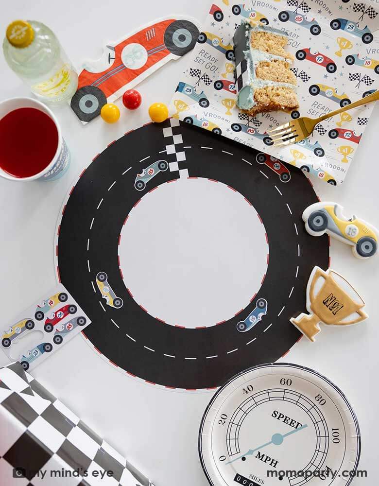 A festive kid's race car themed party table features Momo Party's race track placemats decorated with race car stickers. Around the placemats are race car pattern paper plates, napkins, party cups, speedometer shaped round plates and a checkered table runner by My Mind's Eye. On the table there are some matching race car sugar cookies and a slice of cake, making this a perfect inpo for a car themed bash for your child, be it a TWO FASt 2nd birthday, or a FAST ONE first birthday party.