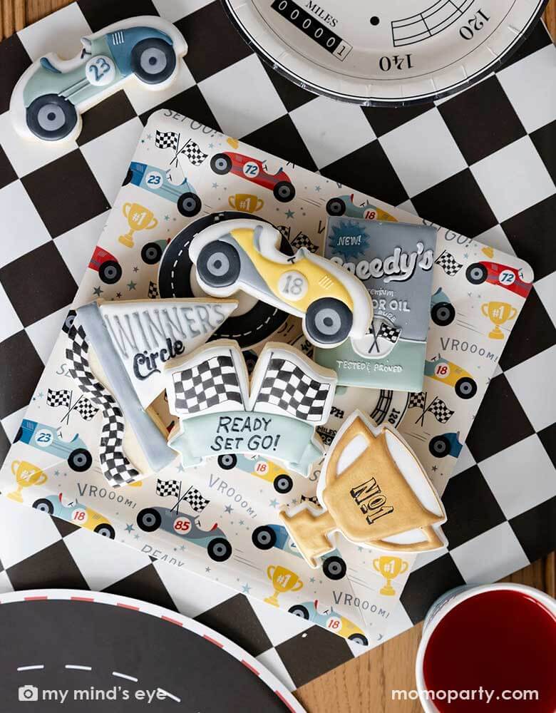 A close-up shot of a race car party table featuring Momo Party's race car pattern square paper plates, speedometer shaped plates and checkered table runner. On the race car pattern plate, there are multiple race car themed sugar cookies with matching style and colors, making this a perfect inspo for kid's race car themed birthday celebration, be it a "Two Fast" second birthday party, a "Fast One" first birthday party.