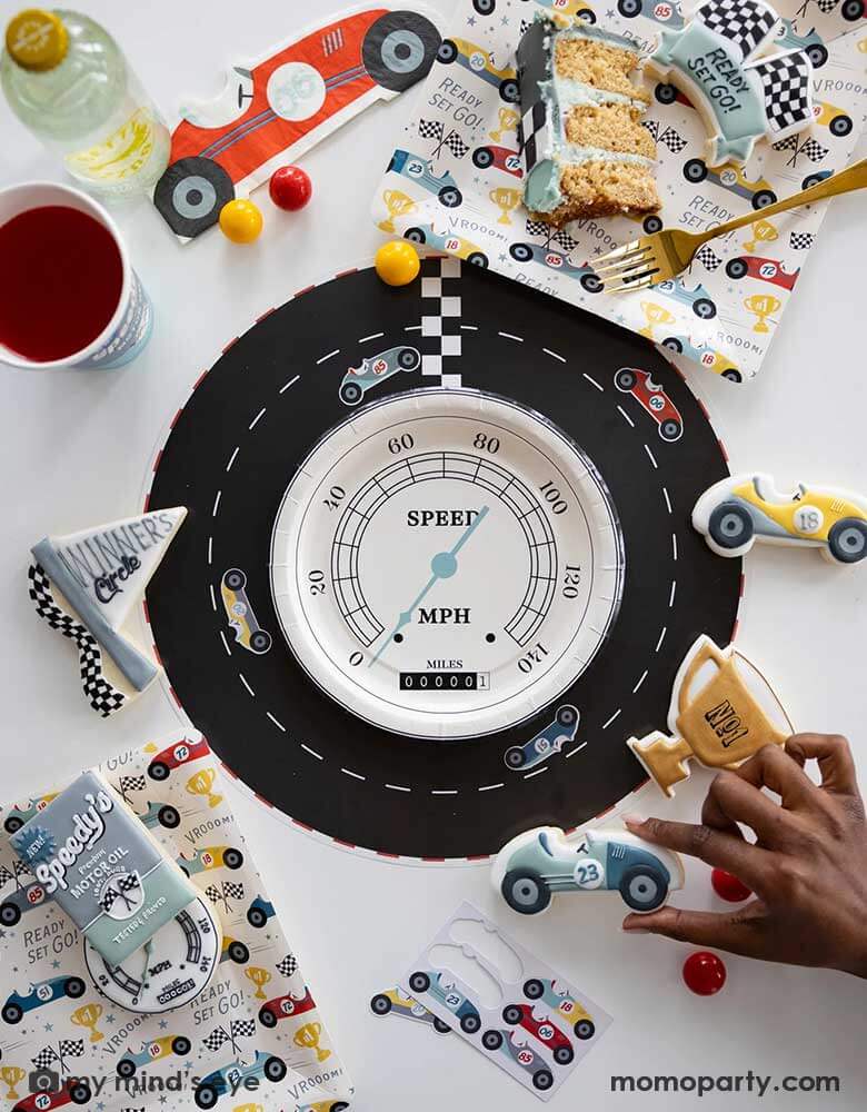 A fun race car themed party table place set featuring adorable race car themed party supplies from Momo Party including a race track placemats adorned with interactive race car stickers in the middle of the placemat is a speedometer shaped plate. Around the tableset laid some race car themed sugar cookies in matching colors and designs around the race car pattern square plates by My Mind's Eye. A perfect inspo for kid's race car themed party tablesetting ideas.