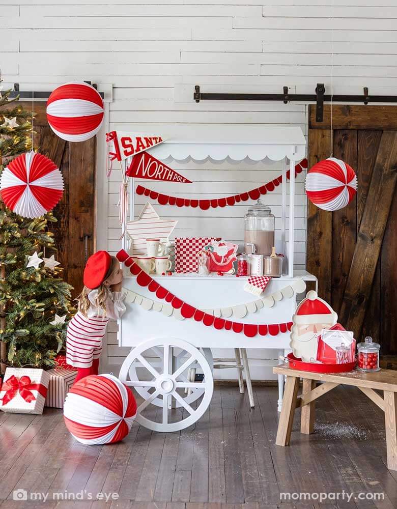 A girl in an adorable Santa themed hot cocoa station set up for a kid's Holiday party featuring a white dessert cart with hot chocolate and Momo Party's Dear Santa party supplies including Santa shaped party cups, checkered plates, napkins, Santa on a sleigh shaped plates and Dear Santa envelope shaped plates. Around the hot chocolate station are two felt party pennant with Santa & Northpole written on them, red scalloped shaped party garlands and some red and white honeycomb balls hung from the ceiling.